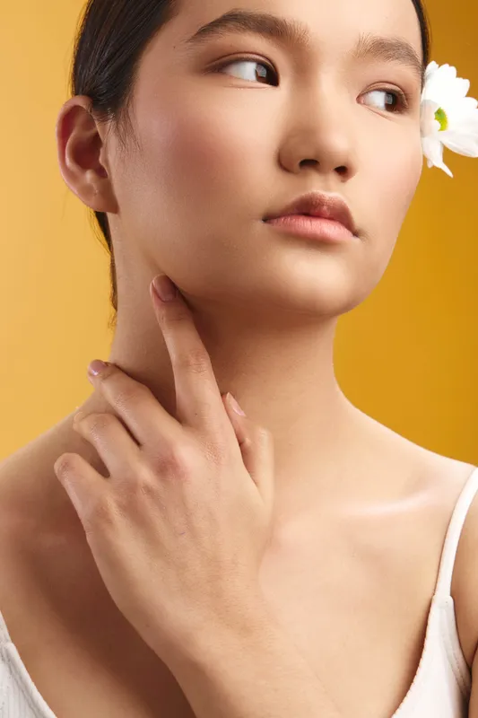 Asian woman is captured in a studio closeup with a flower tucked behind her ear. Soft lighting highlights smooth skin and calm expression, creating a pristine beauty portrait.