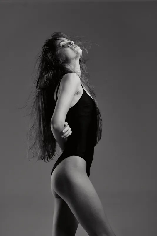 Black and white portrait of a confident woman in a bodysuit posing with closed eyes and long hair against grey background.