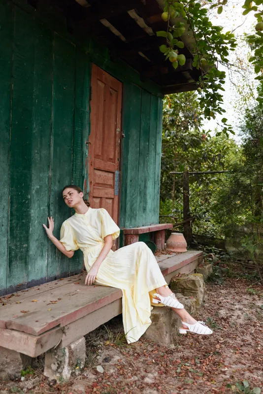 A woman in a pale yellow dress sits on a wooden bench beside a green cottage in a rural outdoor setting, exuding a calm and graceful, peaceful vibe.