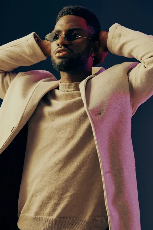 Stylish young Black man in sunglasses, posing confidently with hands behind his head, wearing a light colored sweater and coat against a dark background