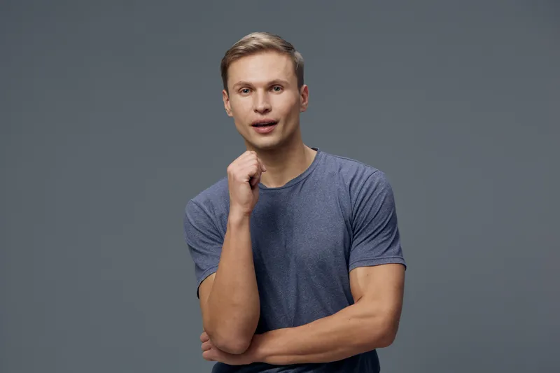 Frustrated young man with casual blue t-shirt and short hair looking worried, holding chin in thought pose, isolated on plain gray background, studio portrait, emotional facial expression.