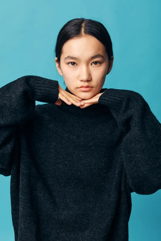 Young woman with black sweater posing with hands under chin on blue background. Portrait of thoughtful female with straight hair wearing casual outfit against solid color backdrop.