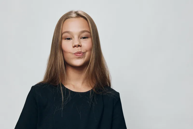 Girl with long hair in black shirt posing and making funny face with lips pursed on gray background in studio. Child expression and casual portrait with natural look and hairstyle.