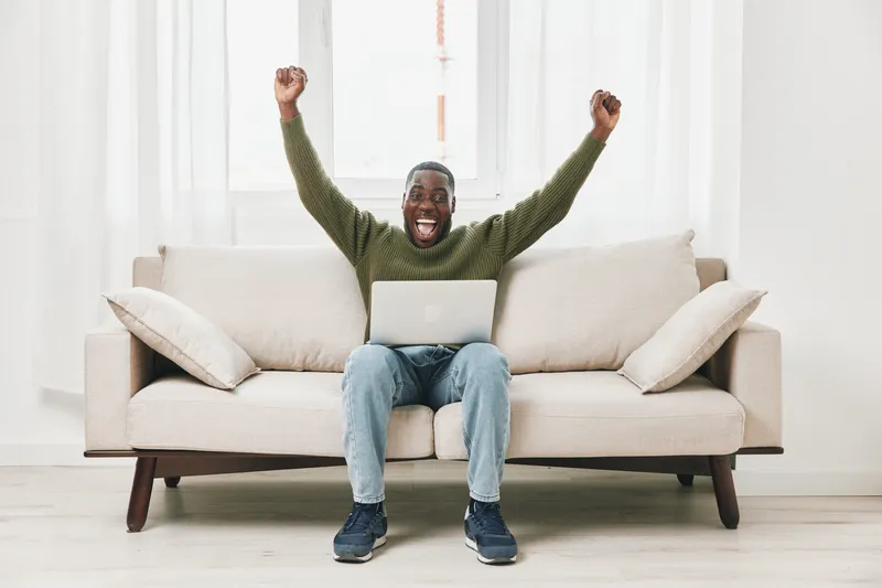 Joyful Black man celebrating success at home with laptop on a cozy couch, wearing a green sweater and jeans, conveying motivation and positivity