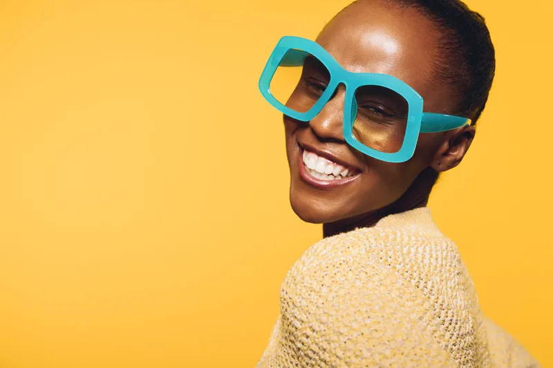 Smiling dark skinned African woman with blue oversized glasses and yellow knitted sweater on yellow background. Concept of fashion, joy, and vibrant style with colorful accessories.