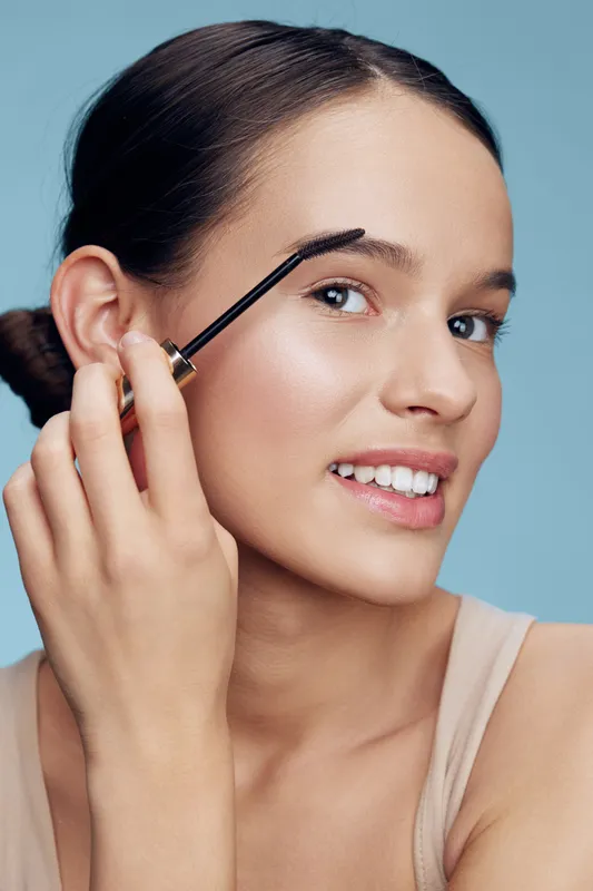 Young woman applying eyebrow gel with a charming smile, featuring a soft skin tone and minimal makeup, against a light blue background for beauty and skincare concepts