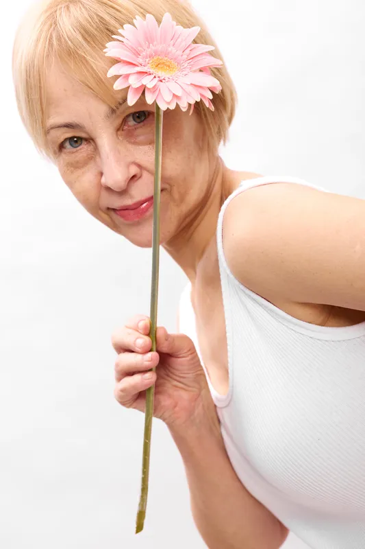 Senior elderly woman, closeup face portrait, holding pink gerbera daisy flower on stem, blonde hair, white sleeveless top, soft studio light, isolated white background