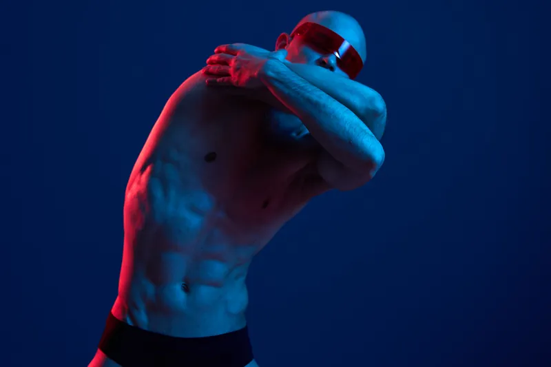 Muscular man posing artistically in a colorful light, showcasing toned physique with vibrant blue and red shades, creating a striking visual impact.