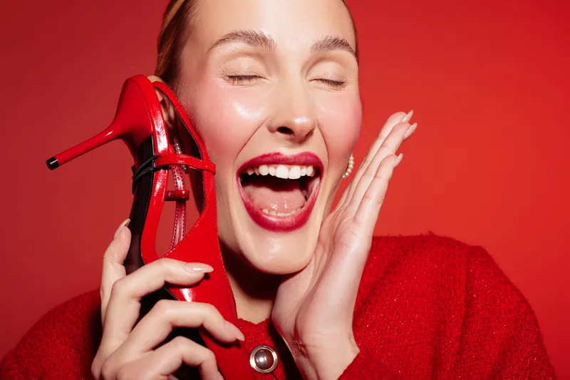 Happy woman with red lipstick and red sweater holds red high heel shoe near face, eyes closed and smiling. Bright red background creates vibrant and joyful fashion concept.