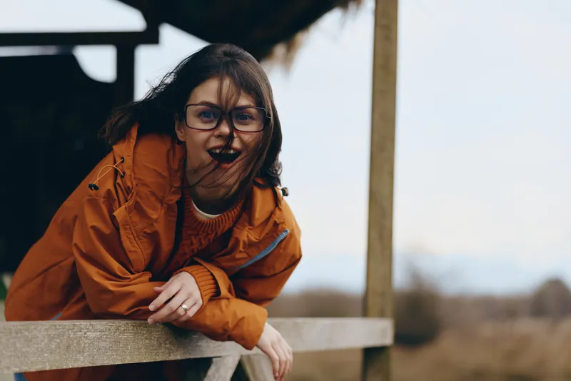 Happy woman lifestyle outdoor portrait smiling with relaxed pose hands resting on wooden railing wearing orange jacket and glasses in garden with soft natural light calm mood today