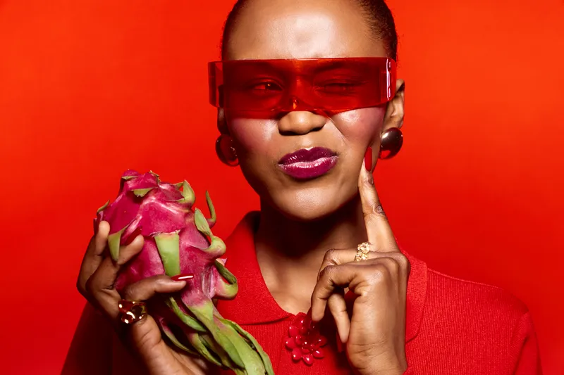 Stylish dark skinned African woman in red sunglasses and clothing holds dragon fruit, posing thoughtfully on red background. Concept for fashion, fruit, and vibrant color contrast.