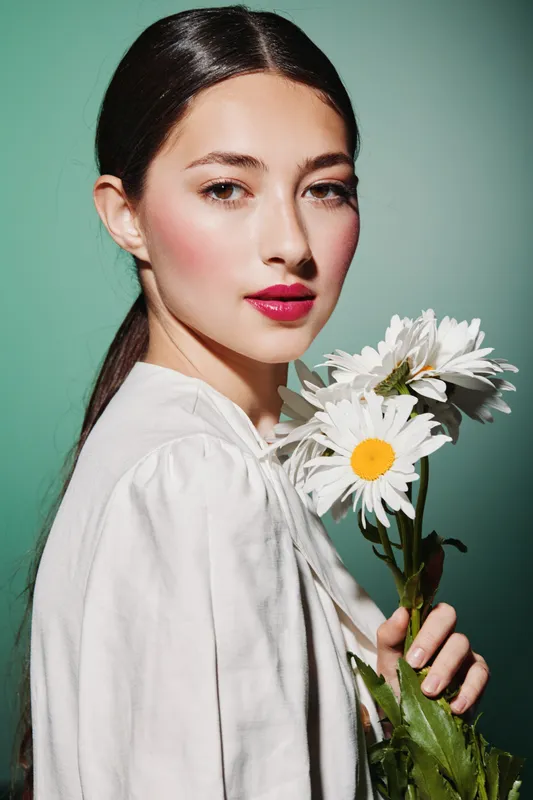 Young woman with sleek dark hair and vibrant pink lipstick holding white daisies. She wears a white blouse against a green background. Concept of beauty fashion and natural elegance.