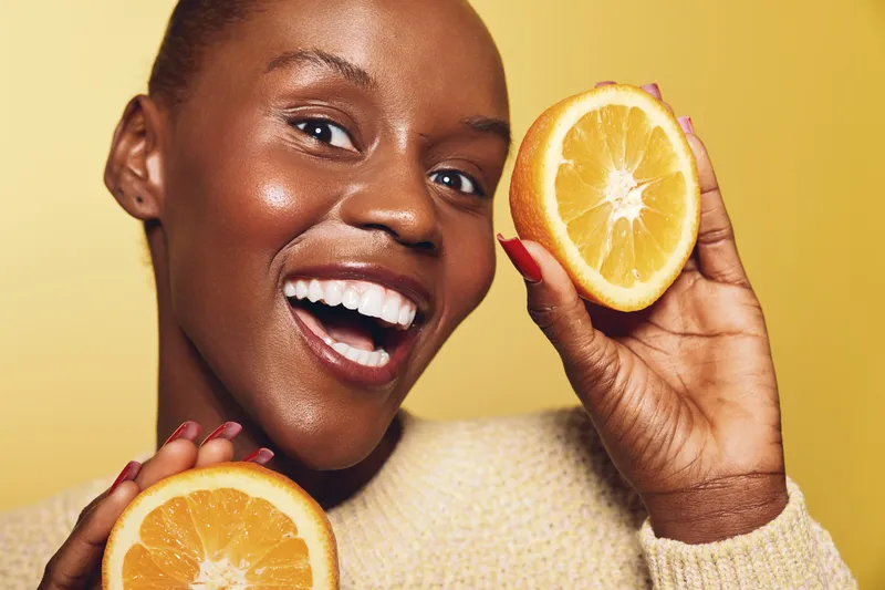 Smiling dark skinned African woman holds two orange halves near face on yellow background. Concept of healthy lifestyle, vitamin C, skincare, and natural beauty with fresh fruit.