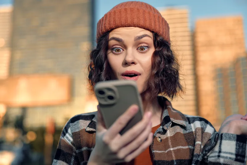 Woman phone surprise in urban outdoors wearing beanie and flannel at golden hour glow, a candid lifestyle scene of authenticity and emotional storytelling capturing mindful living and joy.