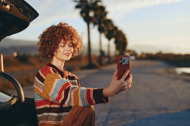 Woman with curly hair takes a selfie with a smartphone near an open car at sunset, striped sweater gives casual travel lifestyle, outdoor travel scene with warm evening light