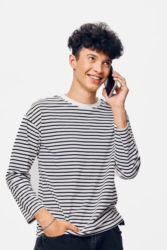 Young man talking on phone with cheerful smile wearing casual striped shirt standing against white background, friendly expression, communication concept.
