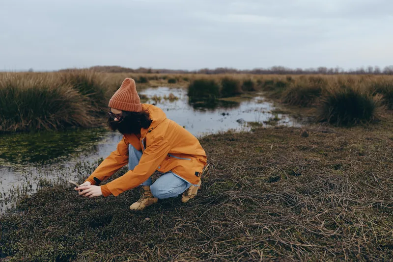 Woman outdoor lifestyle kneels by marsh water, wearing orange jacket with hood, grass and pond reflections in muddy shore under cloudy spring sky, calm nature portrait soft light mood