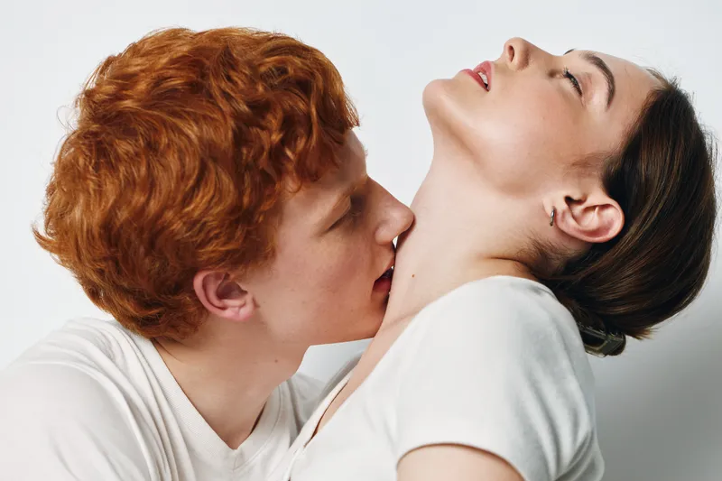 Couple in intimate moment with man kissing woman neck showing affection and closeness, both dressed in simple light clothing against plain background.