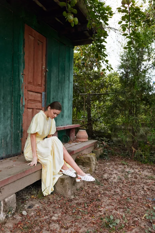 A woman sits on a porch step in a pale dress outdoors in a garden setting, looking down with a calm contemplative mood under leafy shade.
