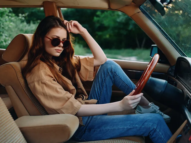 Stylish young woman in sunglasses relaxing in a vintage car, featuring casual clothing, natural emotions, and a lush green background.