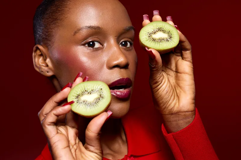 Close-up of dark skinned African woman holding two halves of kiwi fruit near her face on red background. Concept of healthy eating, fresh fruits, beauty, and natural skincare.
