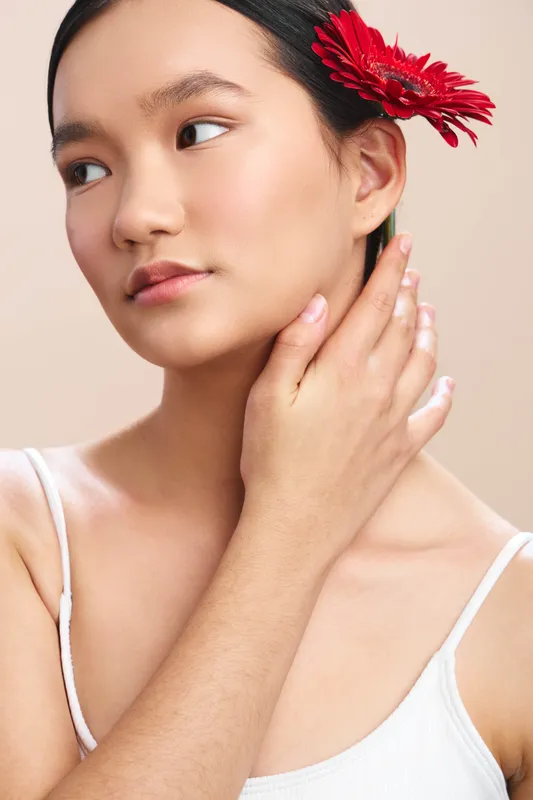 Asian woman touches her neck with a serene expression, posing under soft studio light. A red flower in her hair adds color, creating an elegant, graceful mood and a timeless beauty.
