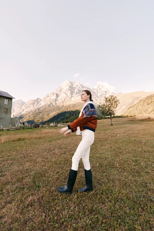 Woman walking in countryside meadow wearing a knit sweater and boots, scenic mountains in background, autumn light, rural cottage nearby, outdoor lifestyle portrait and travel vibe