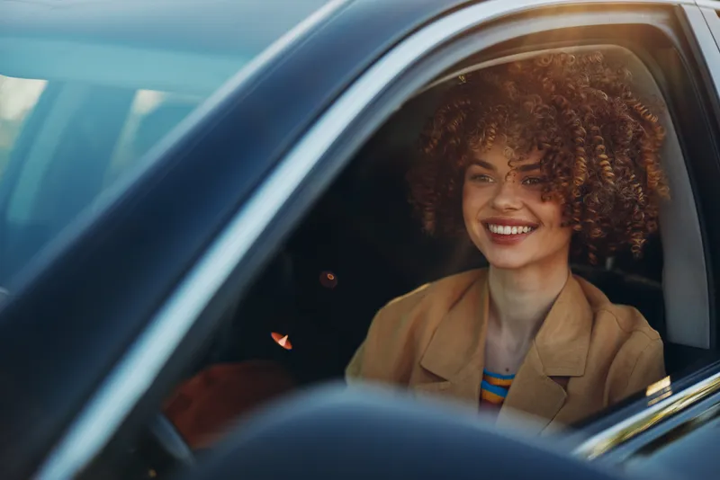Smiling woman with curly hair sitting in a car, daylight on her face, casual jacket, vibrant mood, a candid lifestyle shot emphasizing travel, warmth, and cheerful atmosphere.