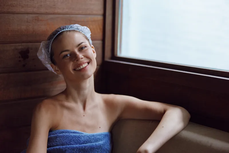 Smiling woman wrapped in blue towel relaxing indoors near wooden wall and large window, enjoying calm, natural light and peaceful atmosphere in spa or sauna setting.