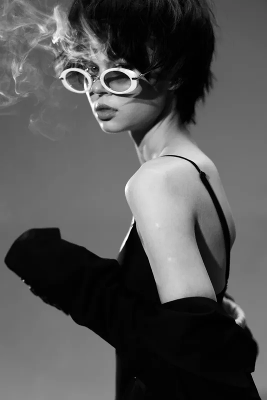 Black and white portrait of a stylish young woman smoking with white sunglasses and a black outfit.