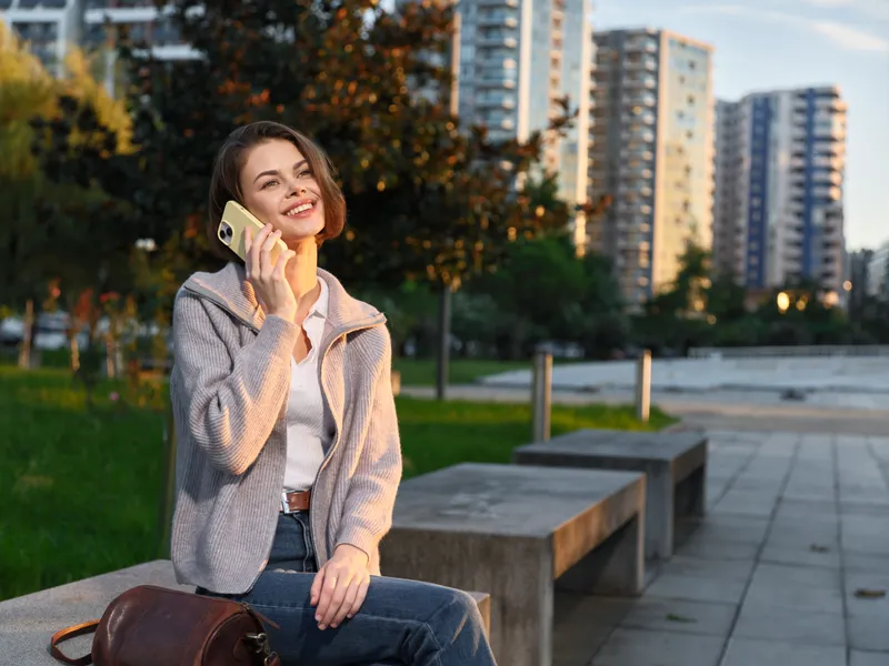 A cheerful woman sits on a park bench in an urban setting, talking on a smartphone. She smiles warmly, relaxed and confident, enjoying a casual outdoor moment amid modern buildings and a calm