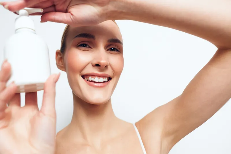 Smiling young woman with clear skin holds a white skincare bottle close to the camera while posing against a bright white background in a beauty and wellness concept.