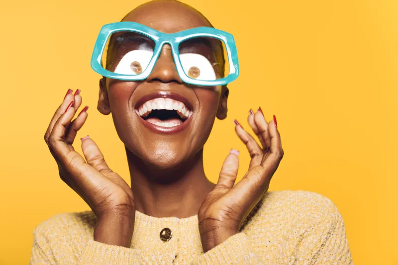 Happy African woman with dark skin wearing large blue sunglasses smiles widely, hands near face. Yellow background creates vibrant, joyful mood. Concept for happiness, fashion, and summer style.