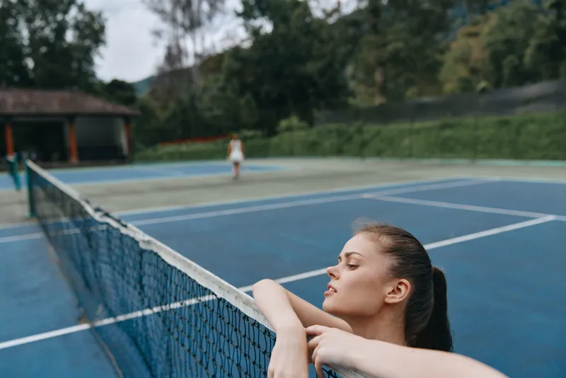Young female tennis player resting by the net, showcasing a focused expression amidst a vibrant green background, perfect for sports and fitness concepts