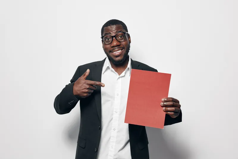 African American man with dark skin wearing glasses and black suit pointing at red paper on white background for presentation or advertisement concept.