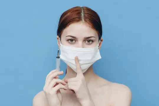 Молодая женщина в медицинской маске с шприцем на голубом фоне