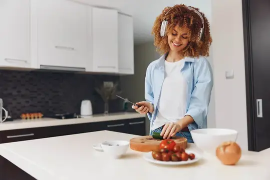 Кудрявая девушка в наушниках режет овощи на светлой кухне