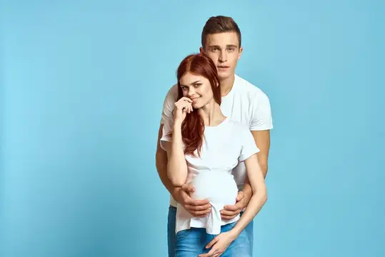 Молодой мужчина обнимает беременную рыжеволосую женщину на голубом фоне