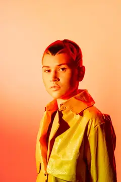 Молодая женщина с заплетенными в косички волосами стоит со спокойным выражением лица, одетая в яркую желтую текстурированную куртку на фоне оранжевого градиента, что подчеркивает современные модные…