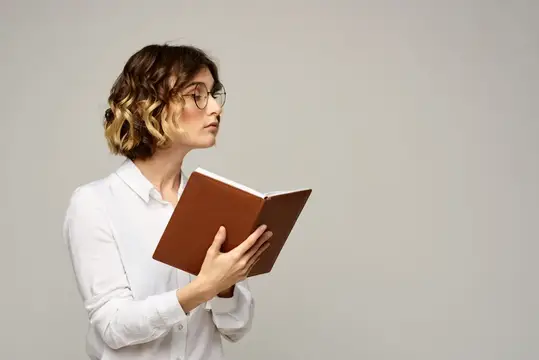 Молодая женщина в очках читает книгу на сером фоне