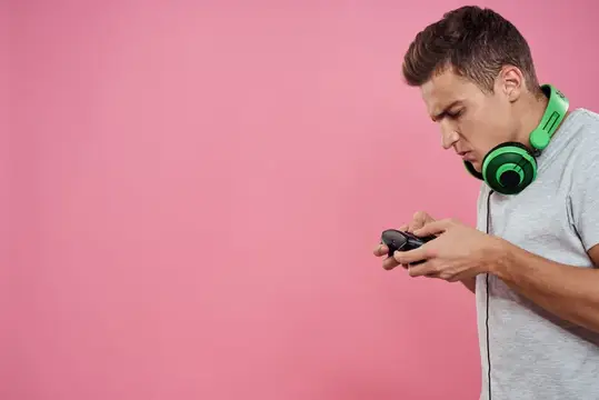 Мужчина с джойстиком в руках и наушниками, играет в игры, развлечения, образ жизни, белая футболка, розовый фон