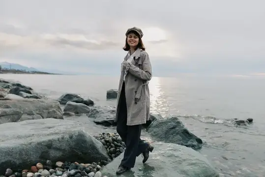Улыбающаяся девушка в плаще и кепке стоит на камнях у моря