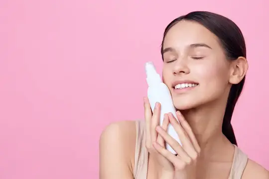 Молодая женщина с закрытыми глазами держит белый флакон на розовом фоне