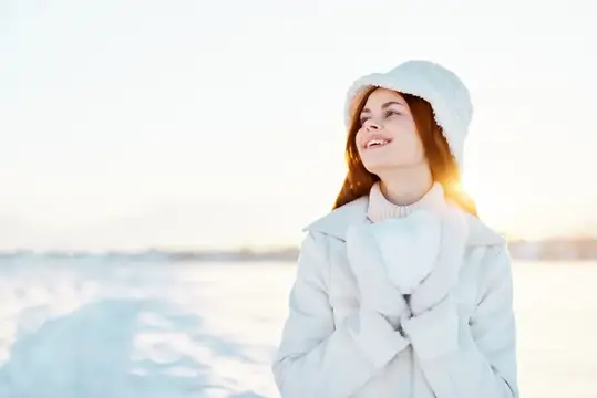 Молодая женщина в белой шапке и пальто улыбается на зимнем снегу