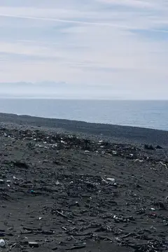 Одинокий прибрежный пейзаж с загрязненным пляжным мусором, темным песком и спокойной водой, далеким горизонтом и туманной атмосферой, подчеркивающий загрязнение окружающей среды и охрану побережья в…