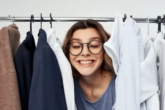 Улыбающаяся девушка в очках выглядывает из-за одежды на вешалках