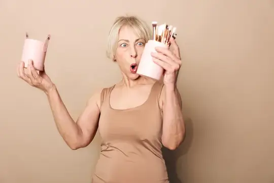 Удивлённая блондинка с кистями для макияжа на бежевом фоне