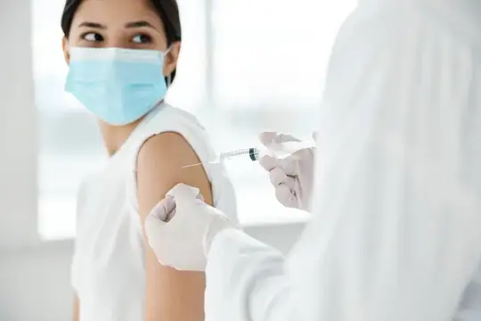 Врач в защитных перчатках делает инъекцию в плечо красивой женщине в медицинской маске