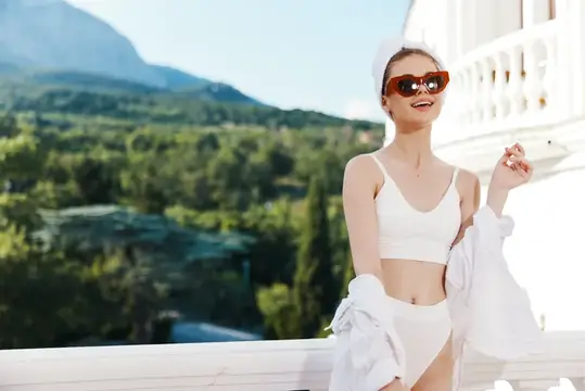 Улыбающаяся девушка в белом купальнике и очках на фоне гор