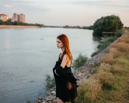 Рыжеволосая женщина в черном платье стоит у реки на природе
